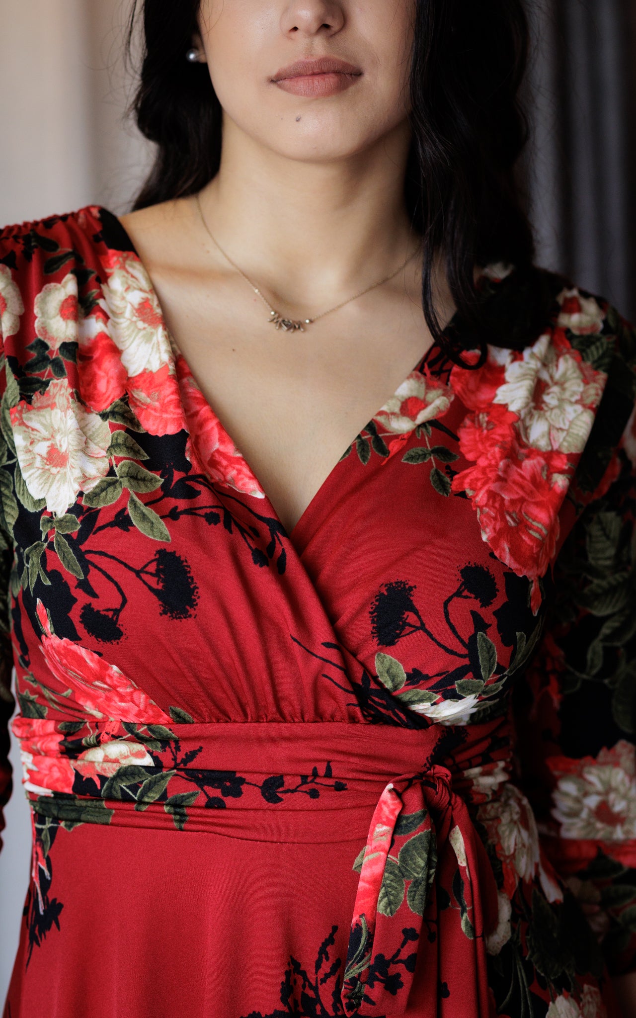 This is a close up of a young lady wearing a V neck style mock wrap dress, with a gathered bust and shoulders and a tie at the waist. The dress is a red floral print. The model has dark hair, worn behind her shoulders, and a dainty gold necklace.