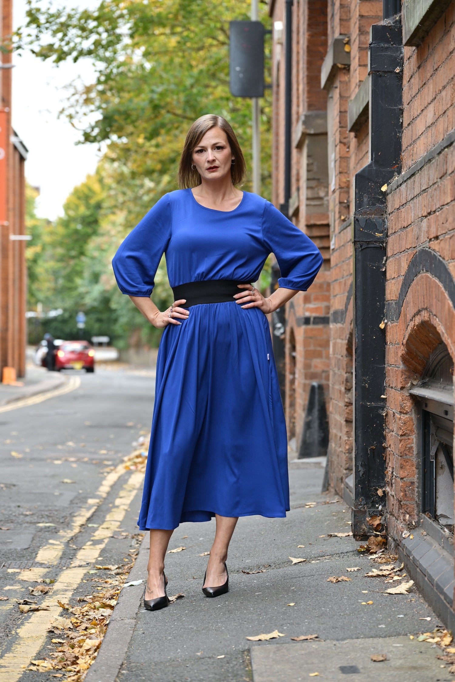 Woman with hands on hips in a blue dress standing on a street with brick buildings.