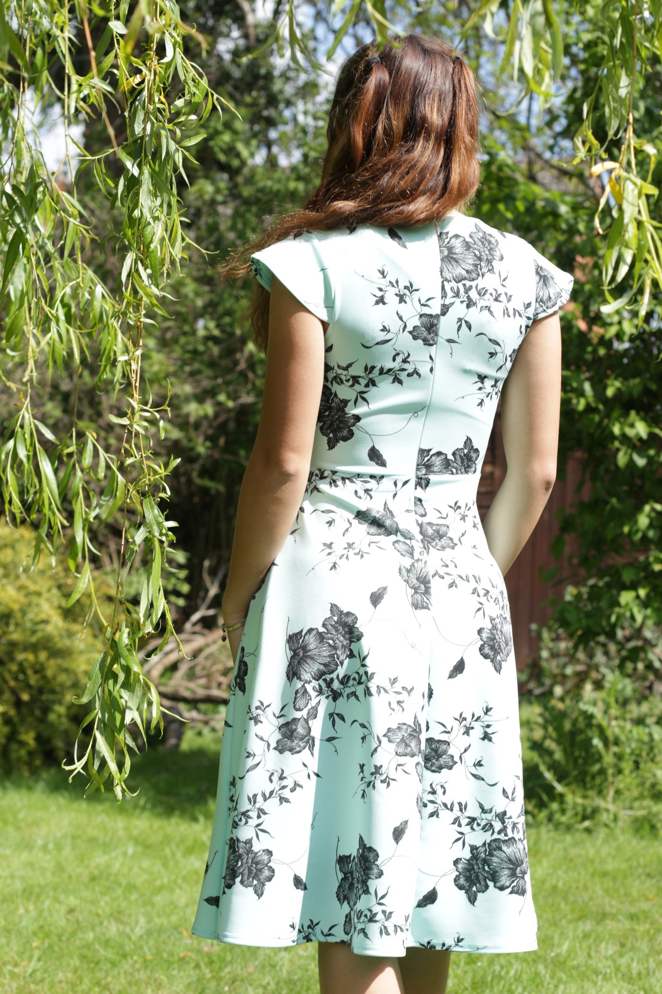 This is the back view of a woman wearing a mint green dress with a black floral print. It has a floaty skirt and short sleeves. The model has brown long hair, swept over her shoulder. She is stood under a willow tree in a garden.