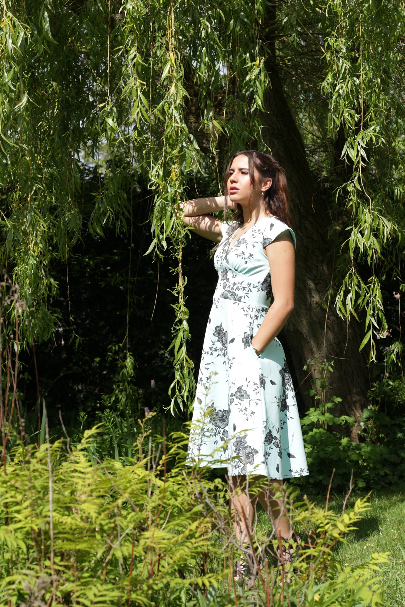 This is a woman wearing skater- style dress in a mint green and black floral print. It has a skirt with lots of movement to it, a high waist and gathered cups below the bust, a V-neck and pockets in the sides. The model has long brown hair and is stood beneath a willow tree in a garden.