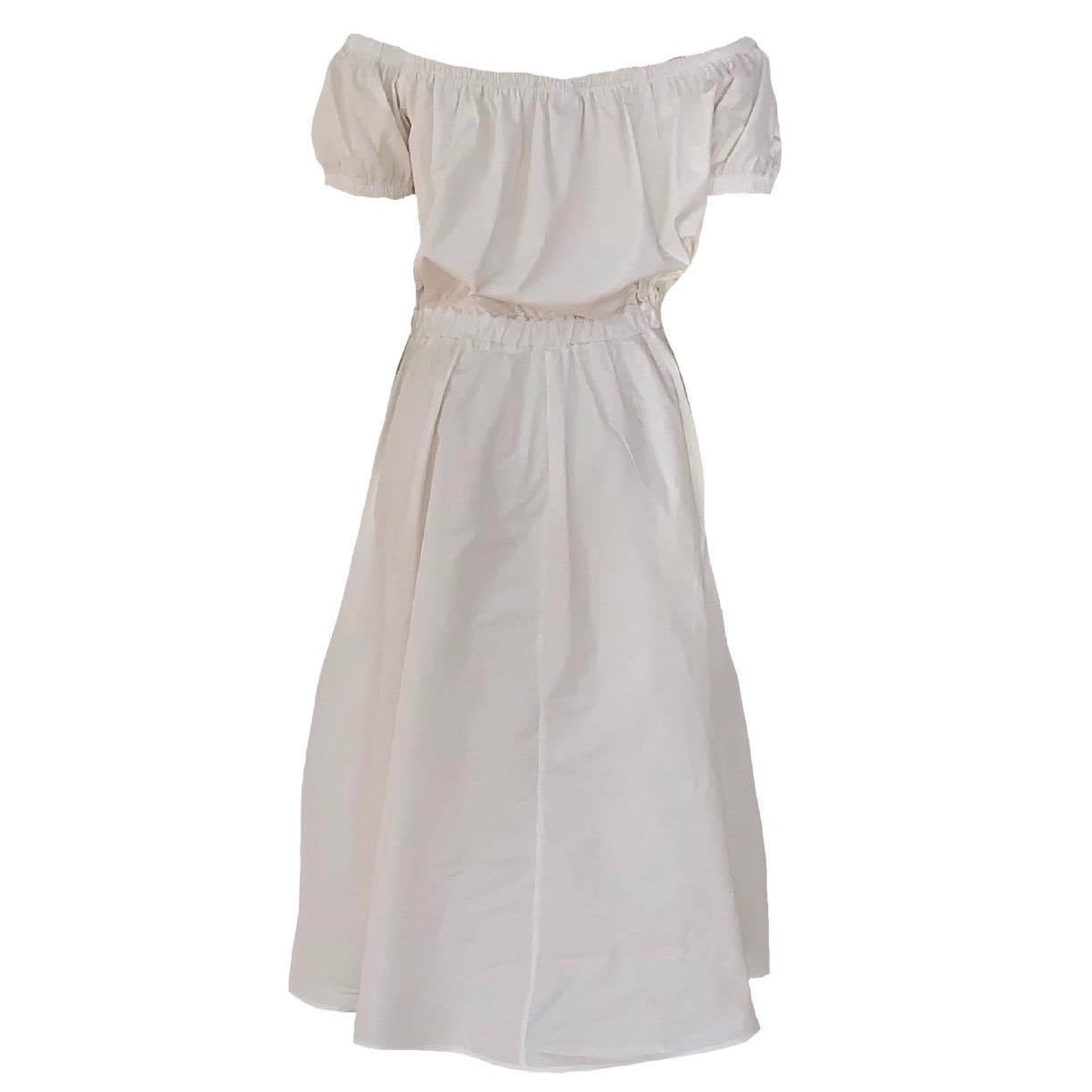 This is a Bardot style dress in cream silhouetted against a white background. the neckline is gathered and the sleeves are a small puff style. The top gathers in to the waist elastic and the skirt is full with side pockets. The dress is a midi length.