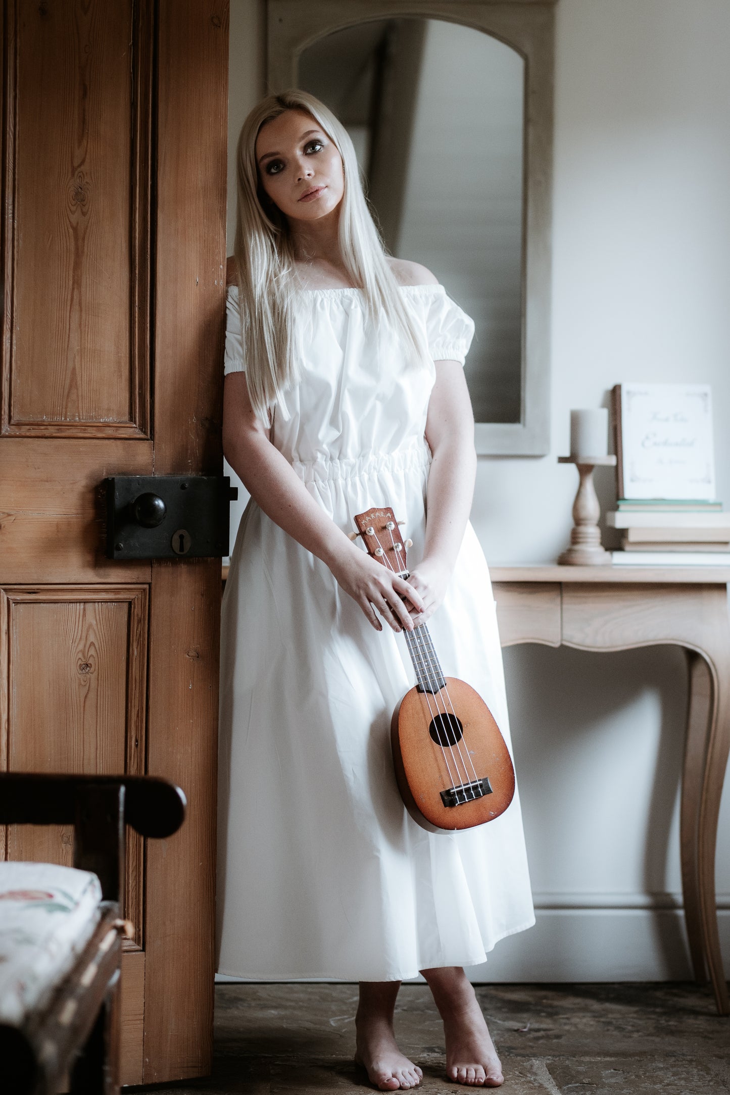 This is a young woman with long blonde hair wearing a Bardot style cream dress with short puff style sleeves. She is silhouetted against an internal room background and is facing the camera leaning against a door. She is holding a guitar and has got bare feet which are crossed.