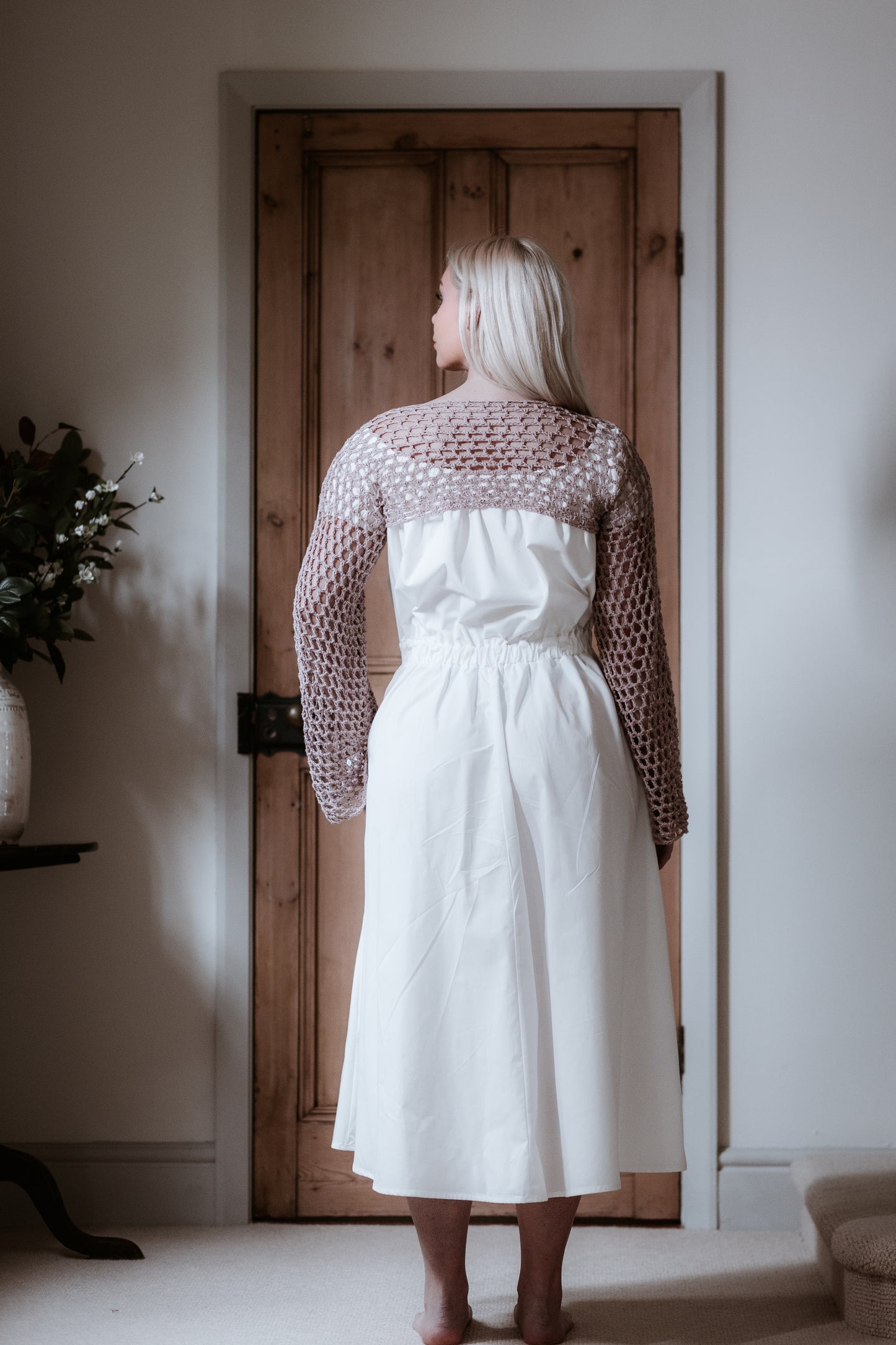 This is a young woman with long blonde hair wearing a Bardot style cream dress with short puff style sleeves. She is silhouetted against an internal room background and has her back to the camera with her head turned to the left. She has her hands at her side and she has got bare feet. She is wearing a pink crochet top over the dress.