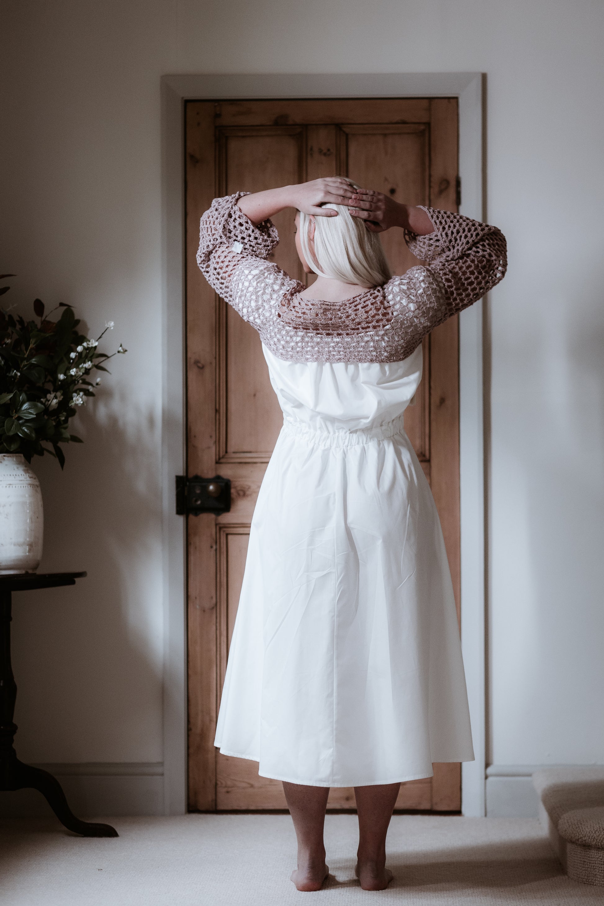 This is a young woman with long blonde hair wearing a Bardot style cream dress with short puff style sleeves. She is silhouetted against an internal room background and has her back to the camera facing a brown wooden door. She has her hands on her head and she has got bare feet. She is wearing a pink crochet top over the dress.