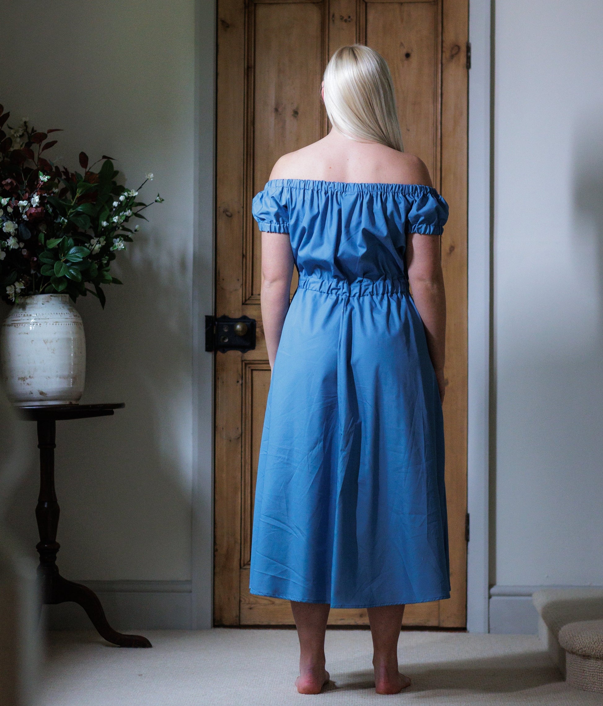 This is a young woman with long blonde hair wearing a Bardot style wedgwood blue dress with short sleeves. She is silhouetted against an internal room background and has her back to the camera facing a wooden door. She has her hands by her side and she has got bare feet.