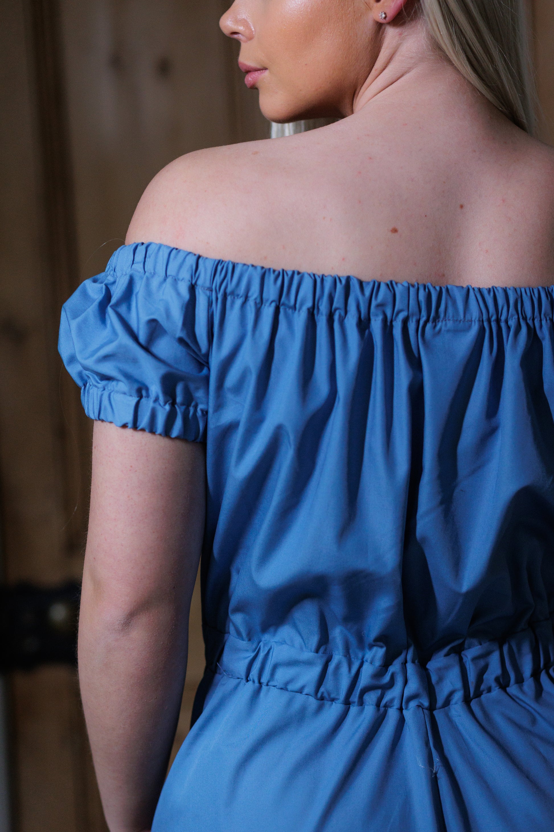 This is a young woman wearing a Bardot style dress in wedgwood blue. This is the top of the dress. She has her back to the camera and her head is turned to the left. The neckline is gathered and shows the left sleeve which is a small puff style. The top gathers in to the waist elastic. 