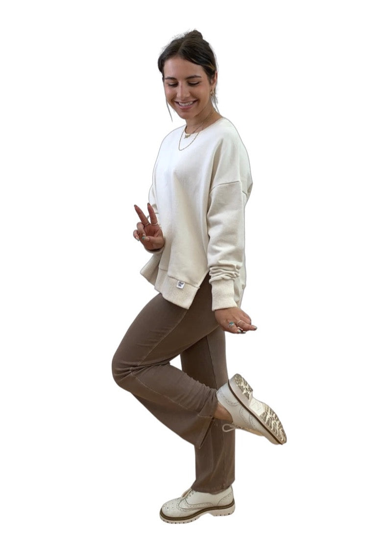 This is a young lady with dark hair. She is silhouetted against a white background and faces the camera. She is wearing  a cream sweat top and mocha trousers. Her right leg is bent at the knee and she is showing the sole of her white shoes.