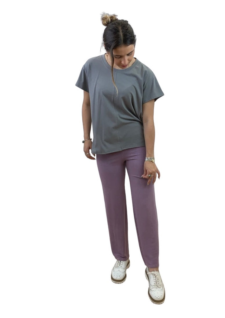 This is a young lady with dark hair wearing a grey T shirt. She is silhouetted against a white background and is looking down at her trousers while she feels the fabric. She is wearing pale grape trousers with white shoes.