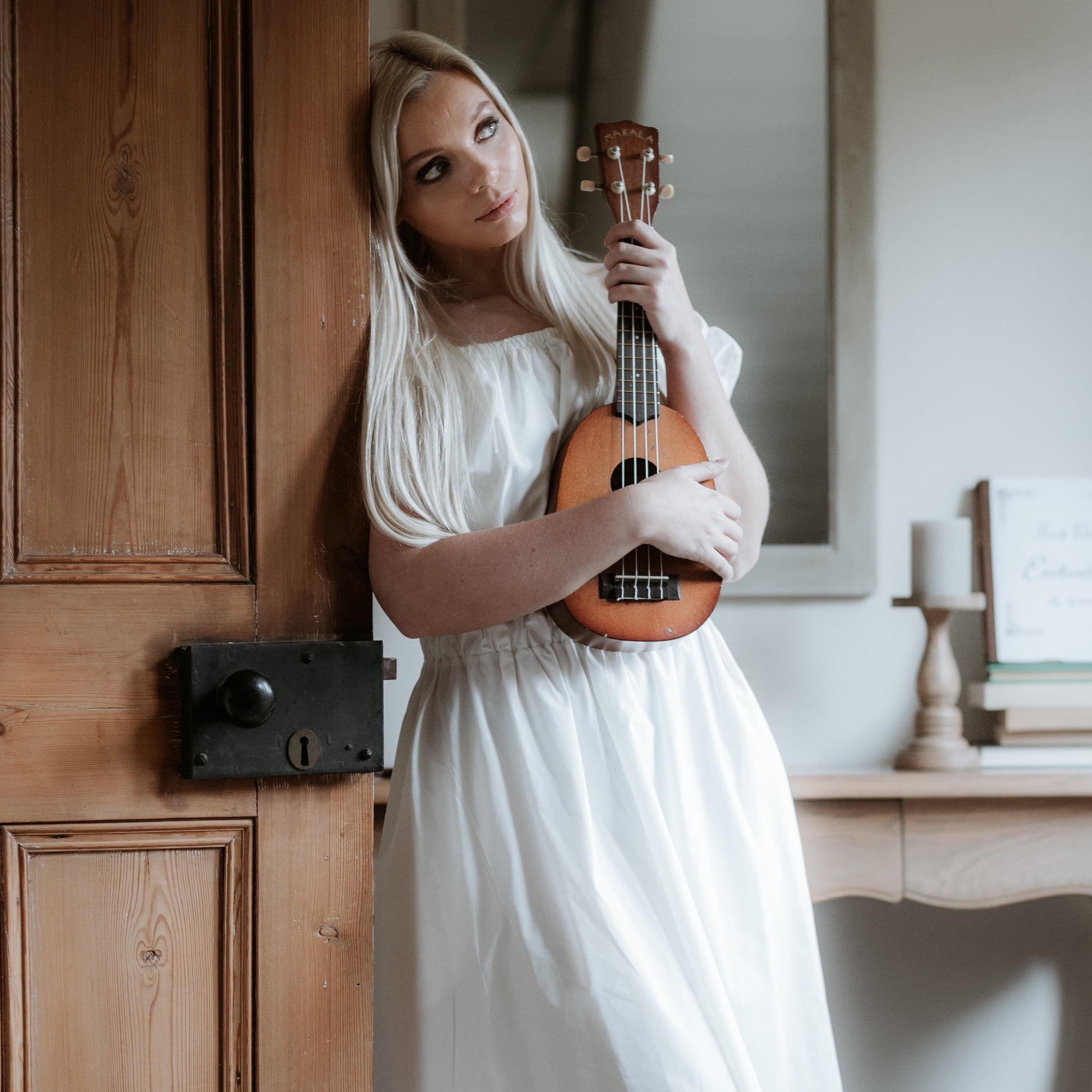 This is a young woman with long blonde hair wearing a Bardot style cream dress with short puff style sleeves. She is silhouetted against an internal room background and is  leaning against a door. She is holding a guitar and looking towards the ceiling.