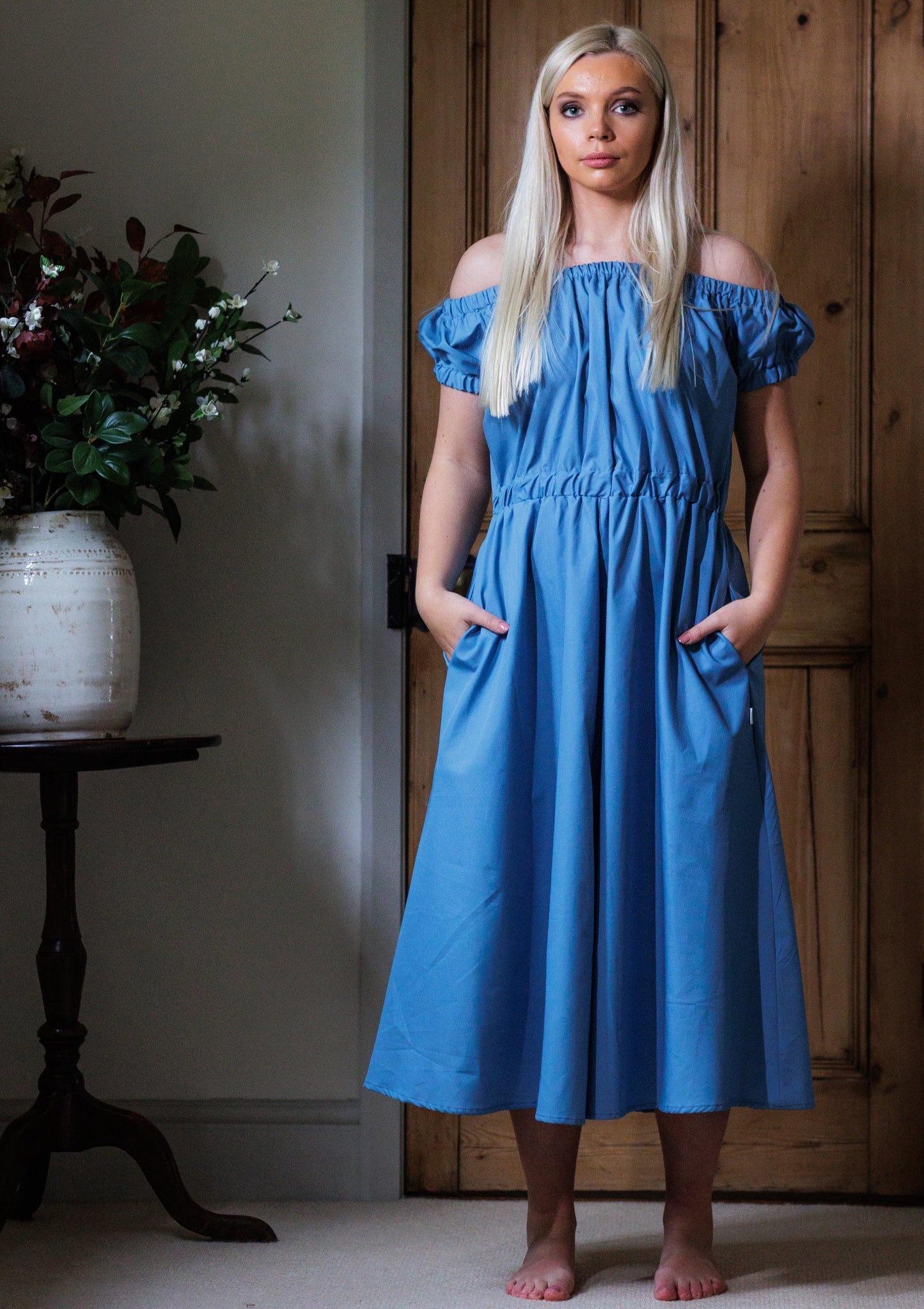 This is a young woman with long blonde hair wearing a Bardot style blue dress with short sleeves. She is silhouetted against an internal room background and is facing the camera. She has her hands in her pockets and she has got bare feet.