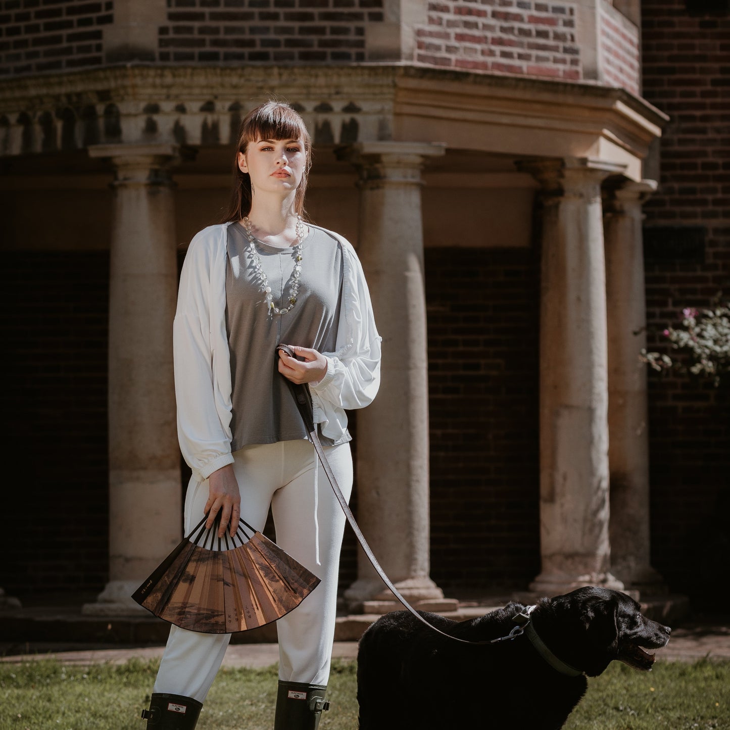 This is a young woman standing in a country garden. She has straight dark hair and is facing the camera. She is holding an open fan in her right hand and she is wearing green wellington boots. She has a black dog on a lead in her left hand. She is wearing a short sleeve tee in steel with a white jacket and trousers.