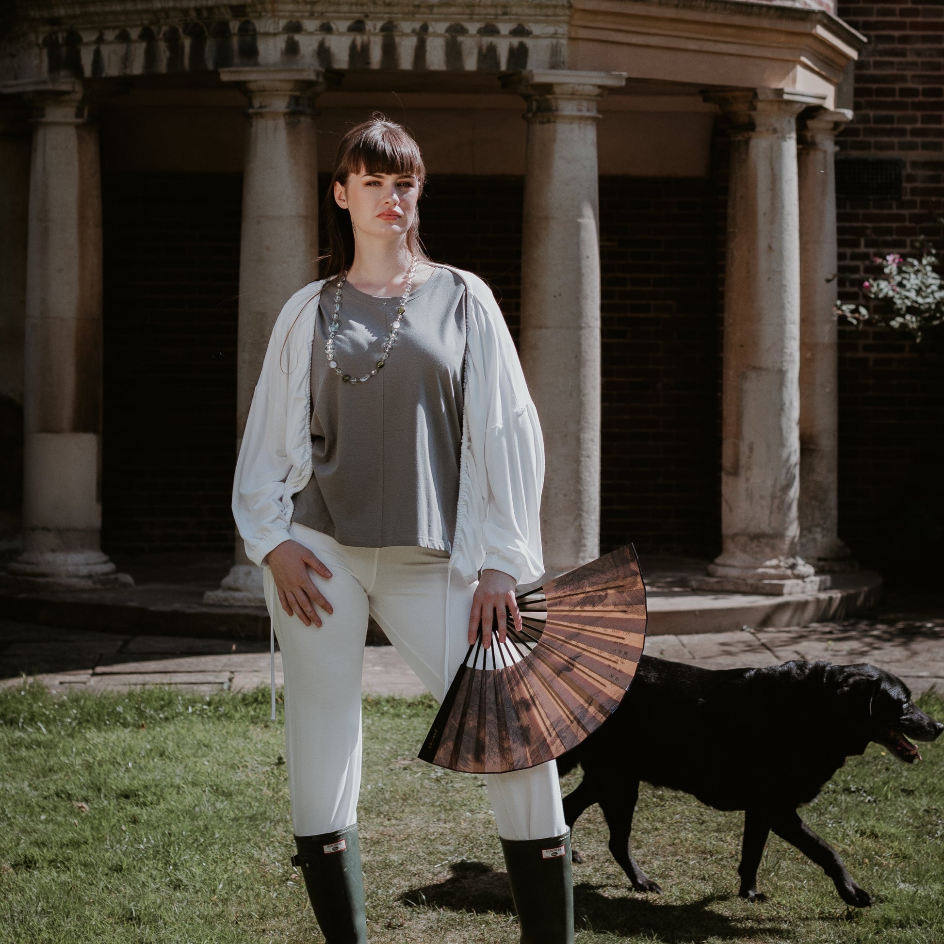 This is a young woman standing in a country garden. She has straight dark hair and is facing the camera. She is holding an open fan in her left hand and she is wearing green wellington boots. There is a black dog at her side. She is wearing a short sleeve tee in steel with a white jacket and trousers.